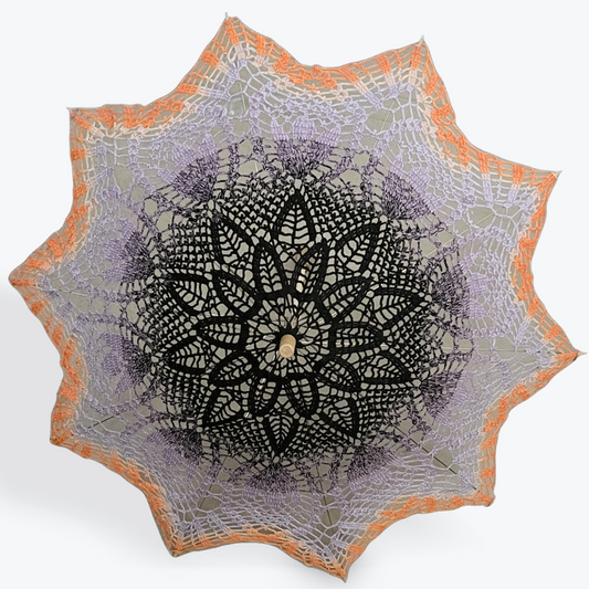 A parasol is open and facing the viewer on a white background. The parasol has a crochet mandala art doily attached which is black at the center and fades into a light lavender purple and then orange at the edge.