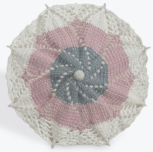 Small decorative parasol is open and facing the viewer. The parasol is blue at the center of the crochet mandala pattern, pink in the middle, and white at the edges. The details on the parasol at the arm tips, center, and handle are all white plastic.