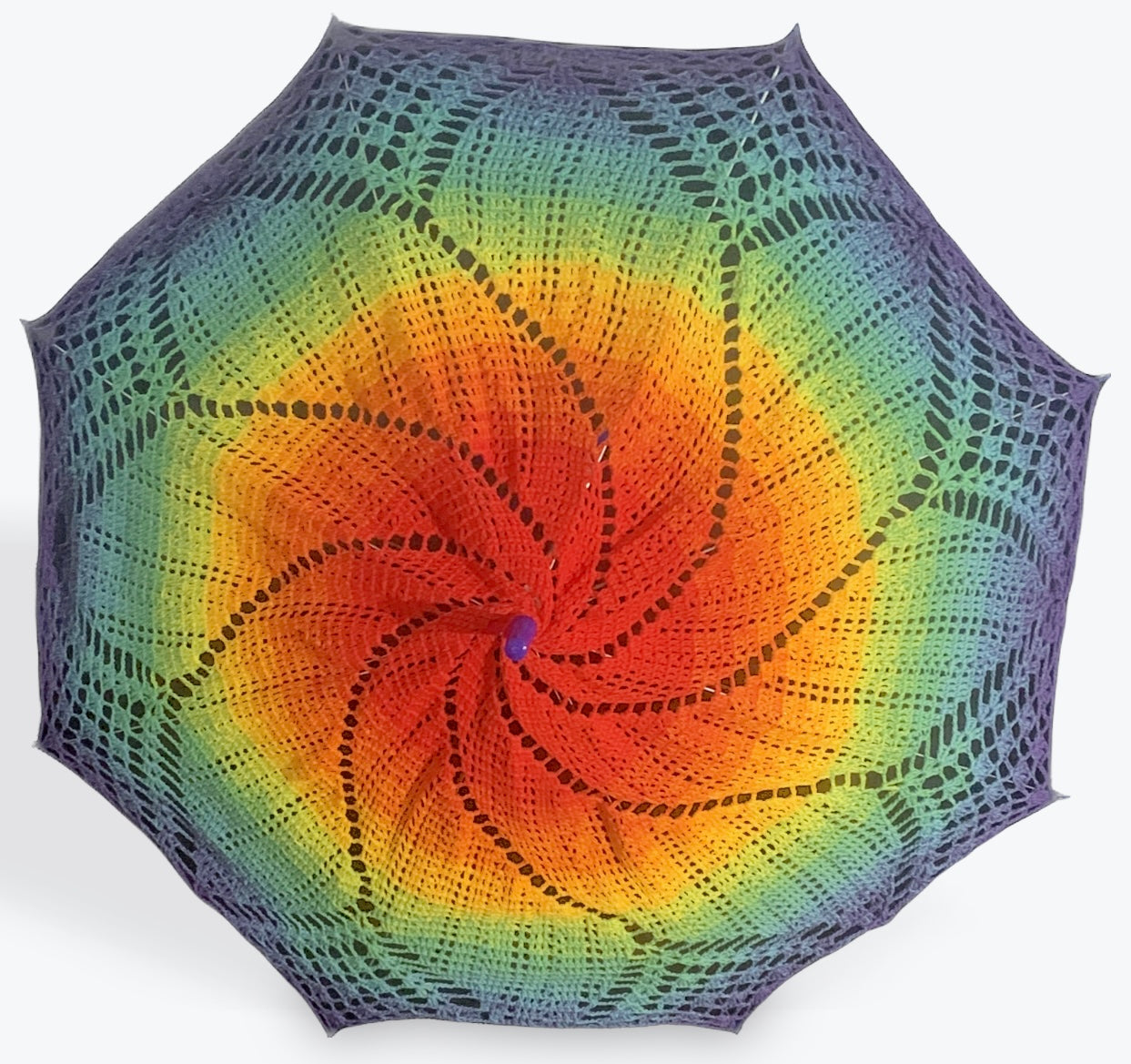 A parasol is open and facing the camera. A crochet mandala adorns this parasol and features the Gay Pride Rainbow Flag in a pattern of 8 petals with small fan sections at the edge.