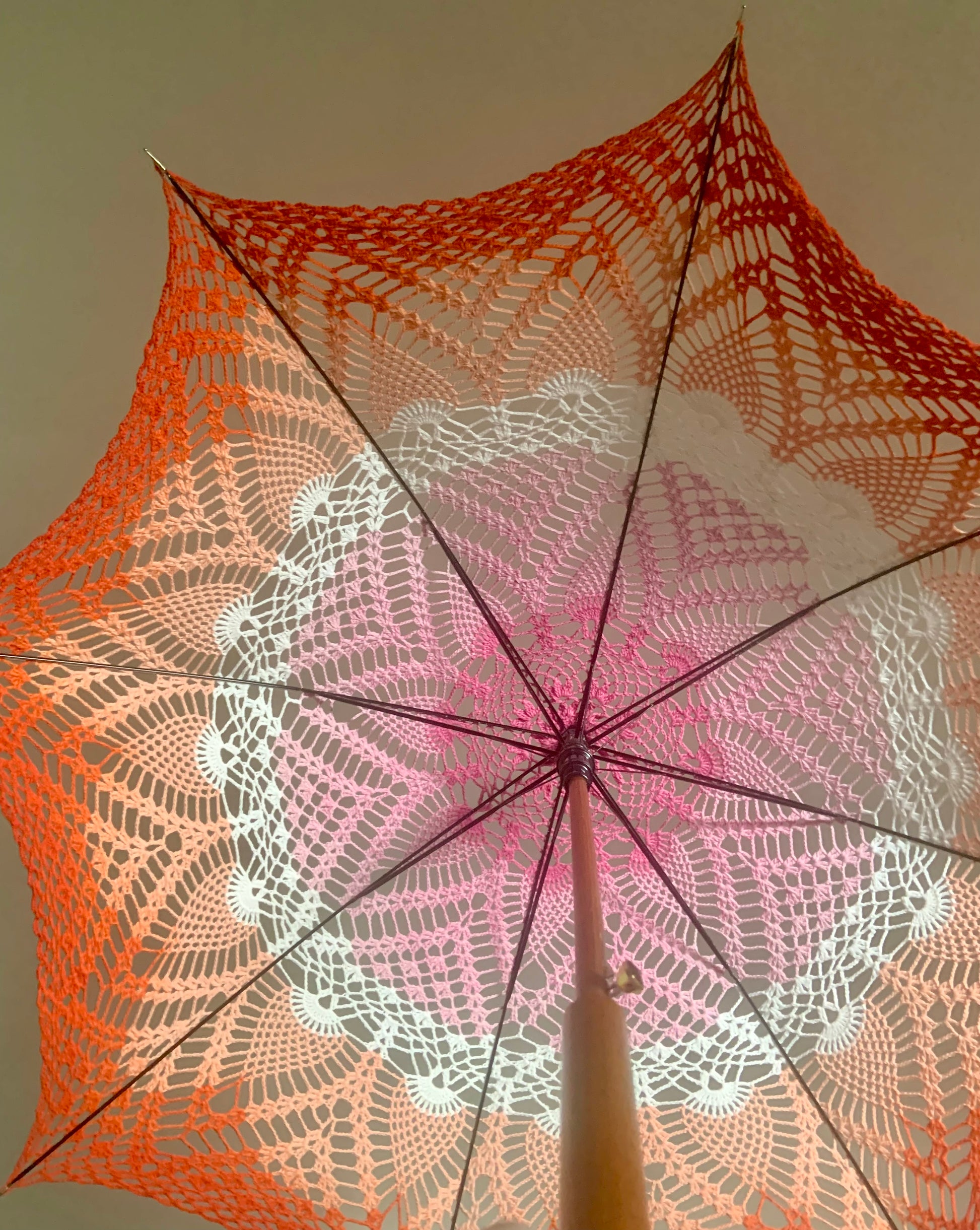 A parasol is open and held aloft over the photographer's head. It features the lesbian pride flag with dark pink at the center followed by light pink, white, light, orange, and dark orange at the edge. The pattern has pineapples and fans throughout. The wooden handle of the parasol extends toward the viewer in the bottom of the photo.