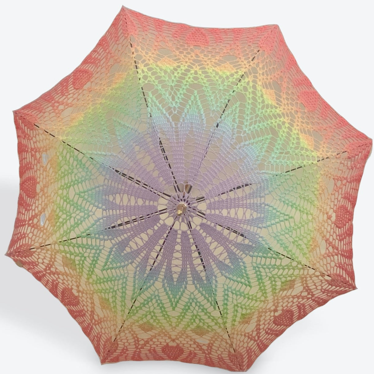 A parasol in a pastel gay pride flag is open and facing the viewer, the black arm are occasionally visible behind the crochet doily.