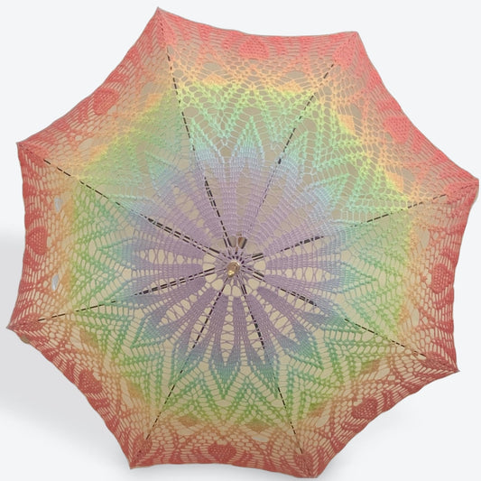 A parasol in a pastel gay pride flag is open and facing the viewer, the black arm are occasionally visible behind the crochet doily.