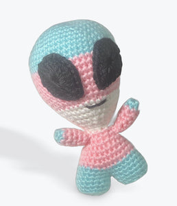 An amigurumi alien with large black eyes, an oval head, small body, arms, and legs fades through the ROYGBIV rainbow like a gay pride flag stands against an endless white background. 