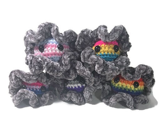 A group of crochet amigurumi oysters are clustered together showing their pearls featuring transgender, pansexual, bisexual, asexual, and gay pride flags. 
