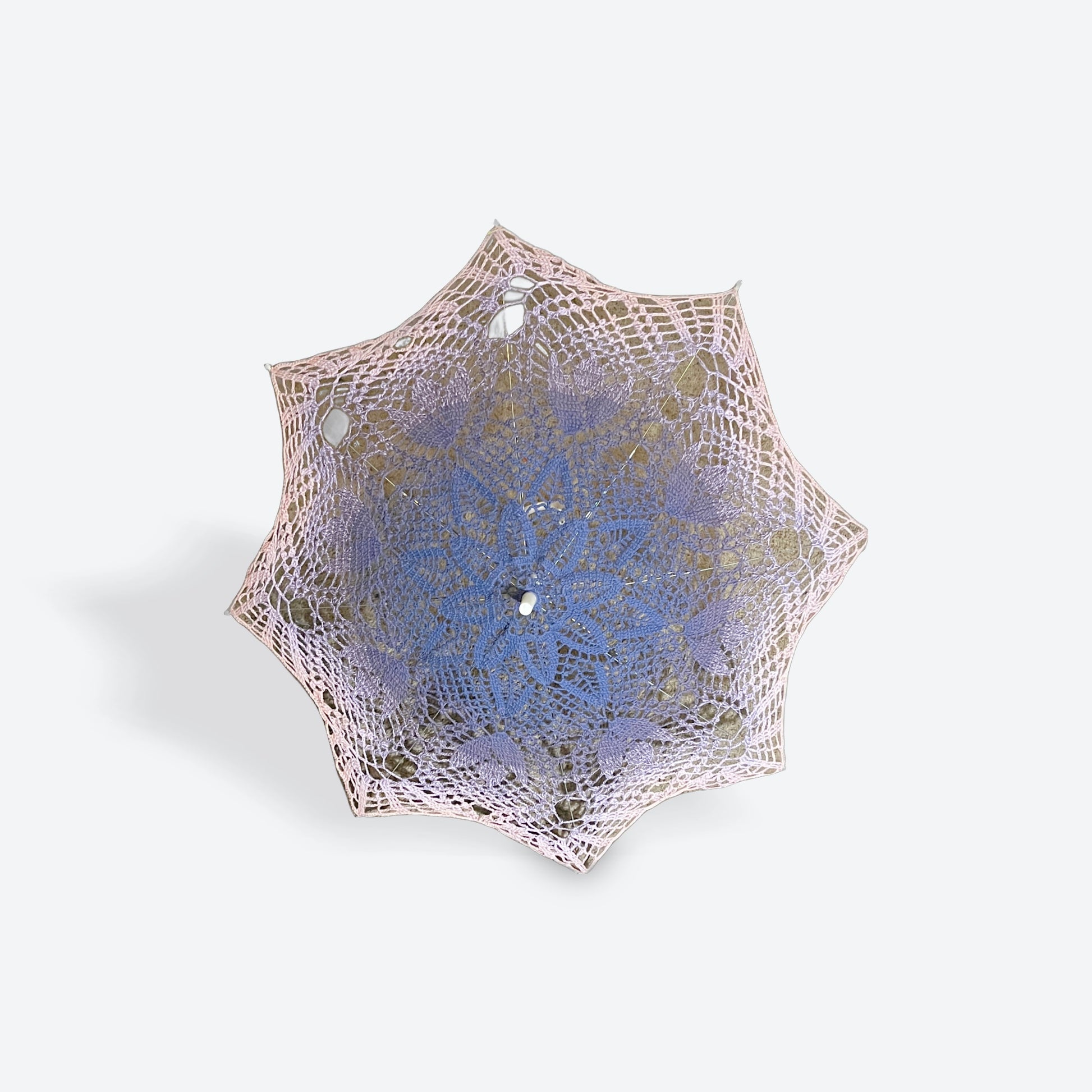 A parasol is open and facing the viewer. This parasol has a fiber art mandala doily attached with begin with purple at the center and fades slowly through light purple to dusty pink at the edge. THe self-drafted pattern has several daisies in the center, a ring of leaves, tulips, and finally fans at the edge. 