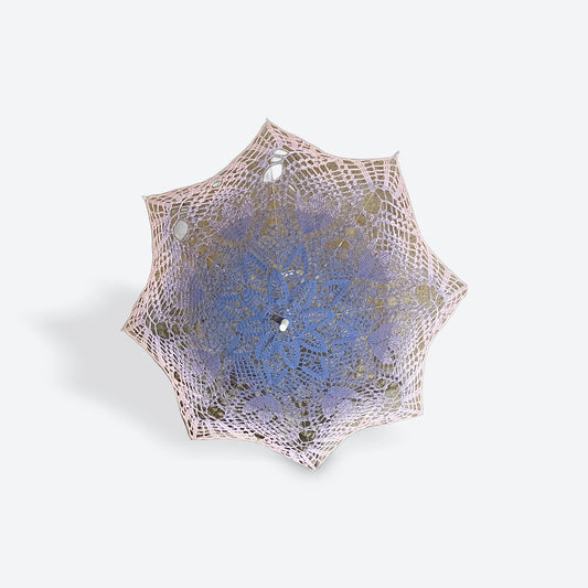 A parasol is open and facing the viewer. This parasol has a fiber art mandala doily attached with begin with purple at the center and fades slowly through light purple to dusty pink at the edge. THe self-drafted pattern has several daisies in the center, a ring of leaves, tulips, and finally fans at the edge. 