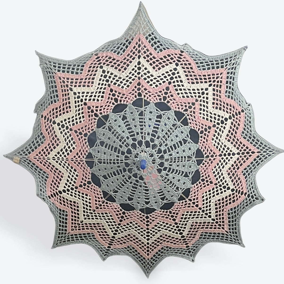 A 30" parasol is open and facing the viewer. A crochet doily has been put on this parasol featuring a chevron patern. The doily features the transgender pride flag with colored stripes going from the center out to the edges in: blue, pink, white, pink, and blue. 