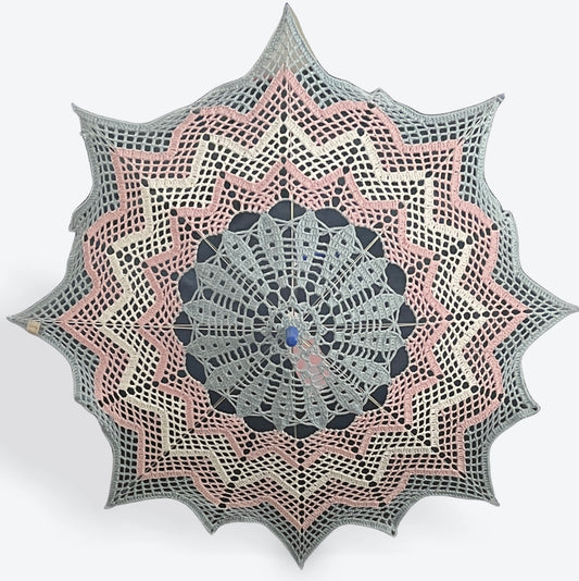 A 30" parasol is open and facing the viewer. A crochet doily has been put on this parasol featuring a chevron patern. The doily features the transgender pride flag with colored stripes going from the center out to the edges in: blue, pink, white, pink, and blue. 