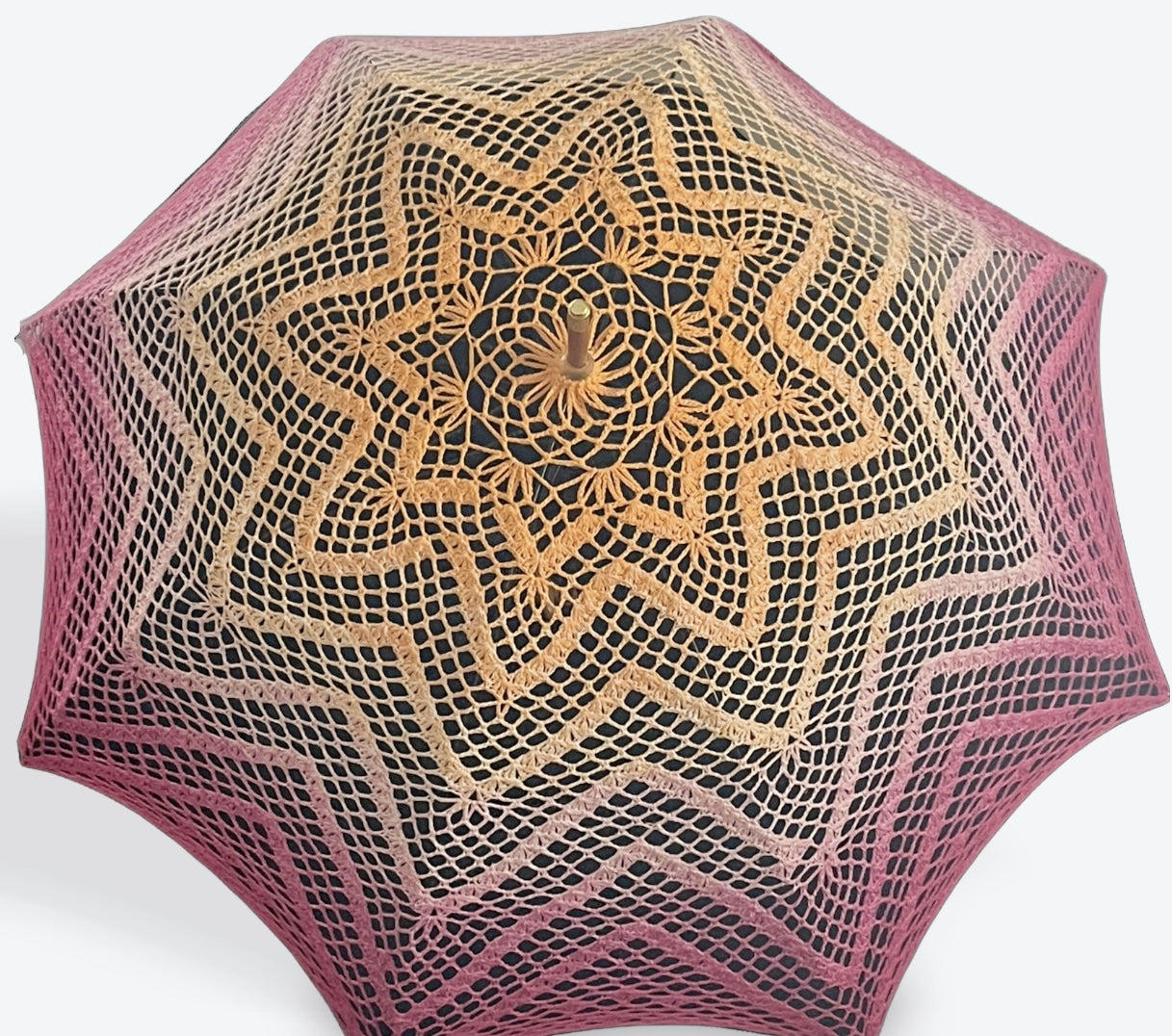 A parasol is open and facing the viewer tilted toward the top of the image. This parasol's cover is hand crocheted in a 100% cotton yarn features the colors of the Lesbian Pride flag. The pattern is 6 repeating concentric 8-pointed stars. 