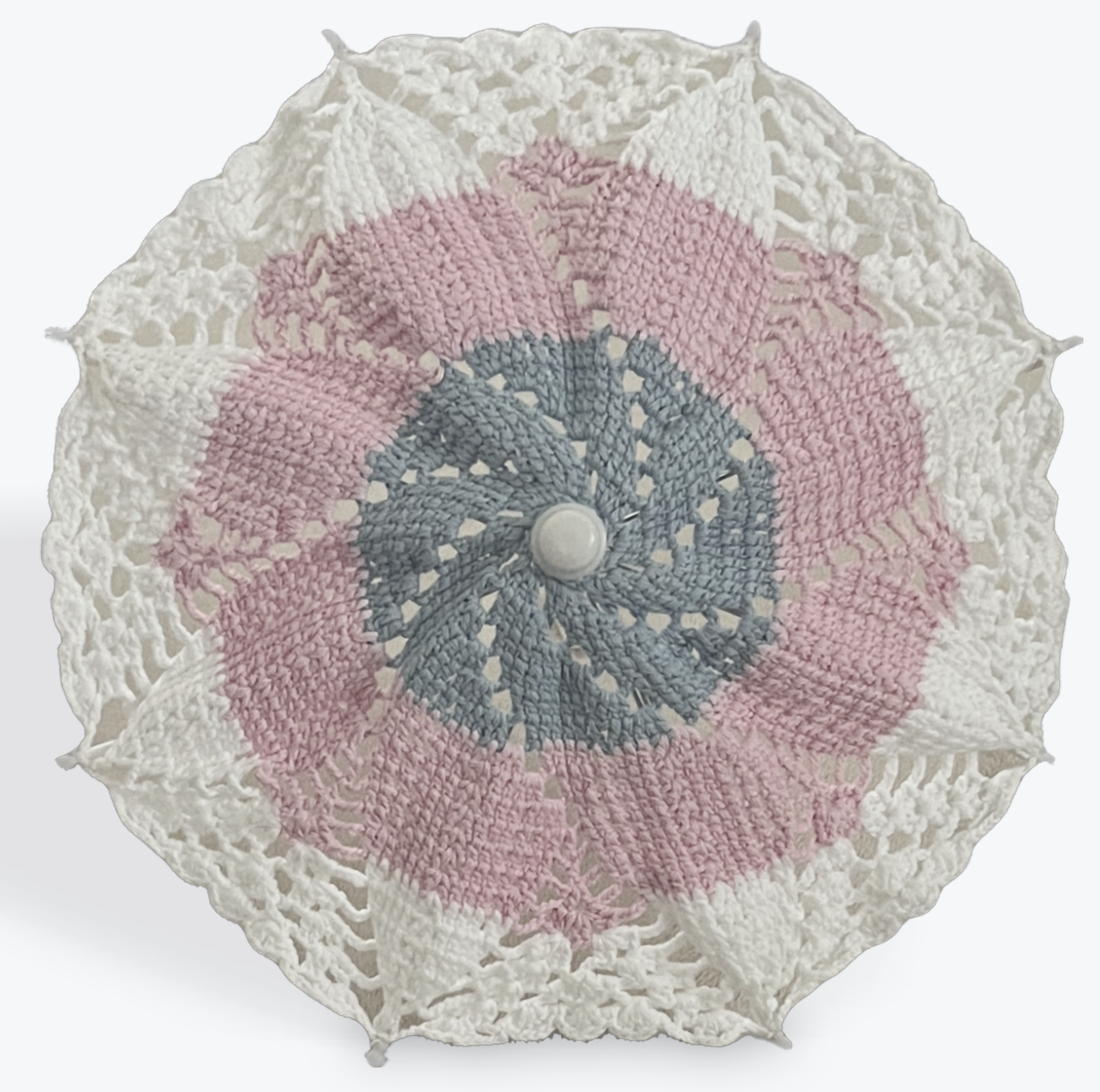 Small decorative parasol is open and facing the viewer. The parasol is blue at the center of the crochet mandala pattern, pink in the middle, and white at the edges. The details on the parasol at the arm tips, center, and handle are all white plastic. 