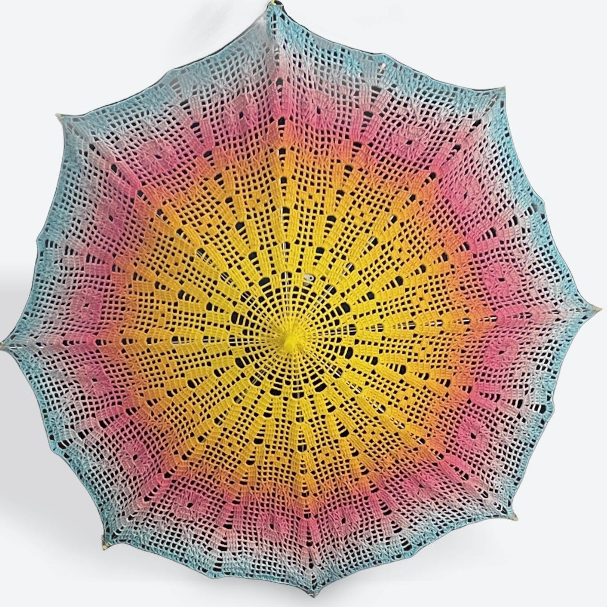 A parasol is open and facing the viewer. It features a filet crochet design. The center is bright yellow and fades into orange, bright pink, soft lavender, and bright blue at the edge. The parasol's center tip is yellow as are the details on each of the 8 arms. 