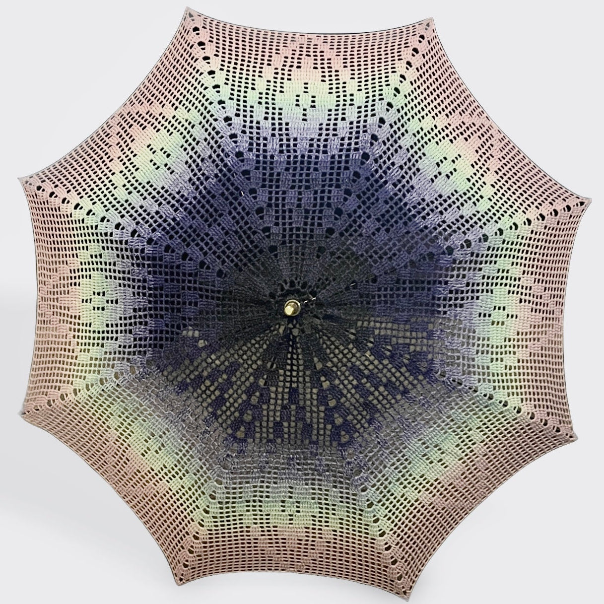 A parasol is open and facing the camera with a filet crochet style doily attached. This doily starts black-blue at the center and fades out to light blue, teal, and pink at the edge. The pattern has a central diamond outlined with a hollow diamond on all 8 panels. 