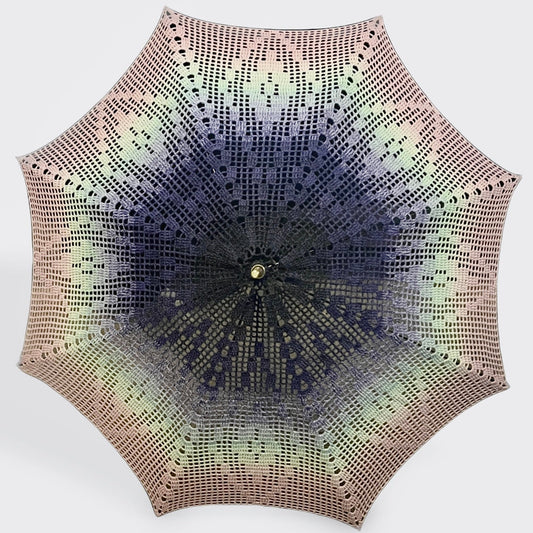 A parasol is open and facing the camera with a filet crochet style doily attached. This doily starts black-blue at the center and fades out to light blue, teal, and pink at the edge. The pattern has a central diamond outlined with a hollow diamond on all 8 panels. 