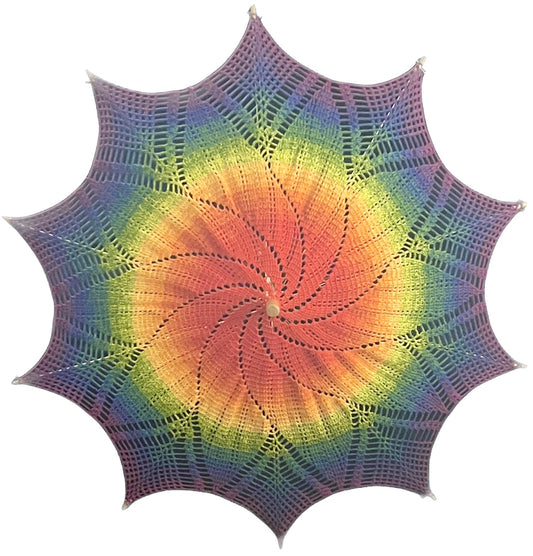 A crochet parasol is open and facing the camera without the handle visible. The mandala art doily on this parasol is rainbow colored with red at the center gradually fading through the ROYGBIV rainbow to purple at the edge. The pattern shows a 10-pointed starburst. 