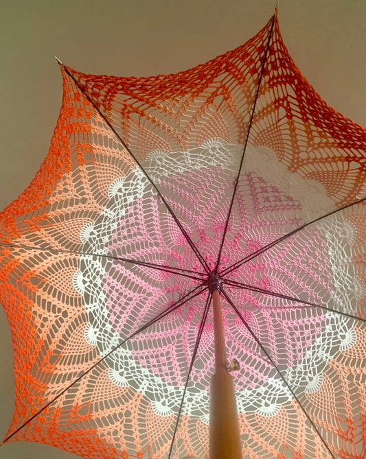 A parasol is open and held aloft over the photographer's head. It features the lesbian pride flag with dark pink at the center followed by light pink, white, light, orange, and dark orange at the edge. The pattern has pineapples and fans throughout. The wooden handle of the parasol extends toward the viewer in the bottom of the photo. 