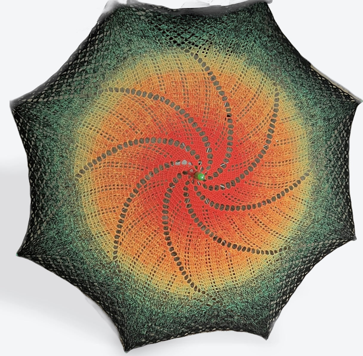 A parasol is open and facing the viewer. This parasol's cover is crocheted in a swirling pattern with lots of sun coverage. The parasol is dark orange at the center fading slowly to yellow, light green, dark green, and finally black at the edges. The arm tips and topper are green plastic. 