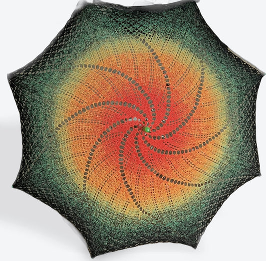 A parasol is open and facing the viewer. This parasol's cover is crocheted in a swirling pattern with lots of sun coverage. The parasol is dark orange at the center fading slowly to yellow, light green, dark green, and finally black at the edges. The arm tips and topper are green plastic. 