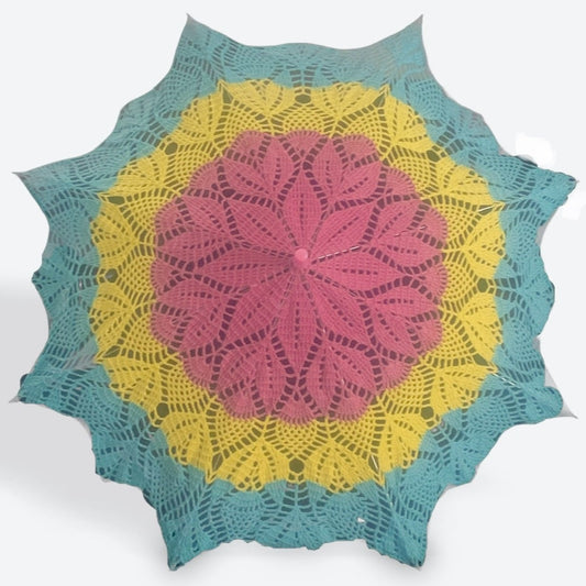 A 30" parasol is open facing the camera. The doily attached to the parasol is pink at the center followed by yellow and blue at the edge. A kaleidoscopic pattern of pineapples and leaves is present throughout. The top and arm tips are light pink.  