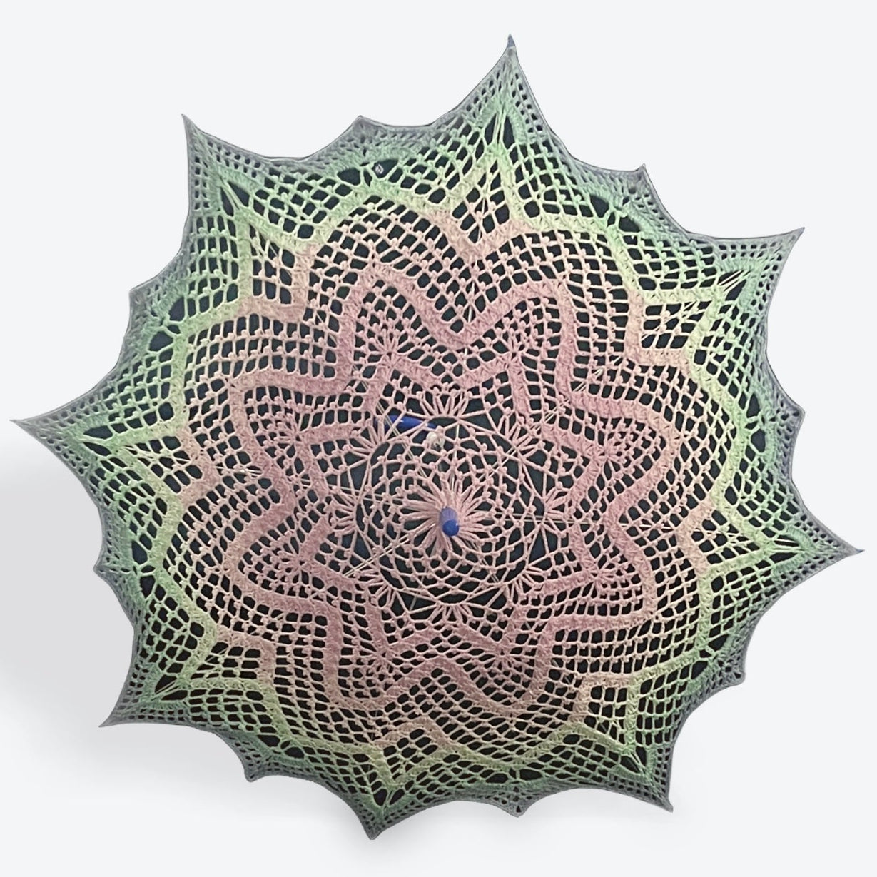 A parasol is open and facing the viewer. Its cover is a crochet fiber art doily featuring an 8-pointed star repeated concentrically 3 times with sharp spikes added on the last round to make 16 points around the outer edge. The parasol fades gradiently from the center pink, then teal, then light blue at the edge. 