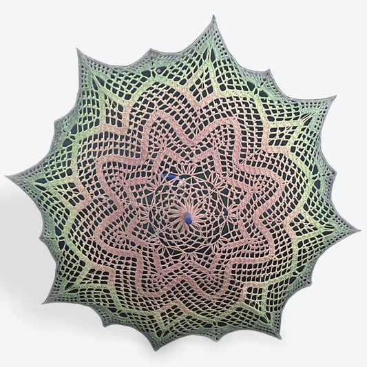 A parasol is open and facing the viewer. Its cover is a crochet fiber art doily featuring an 8-pointed star repeated concentrically 3 times with sharp spikes added on the last round to make 16 points around the outer edge. The parasol fades gradiently from the center pink, then teal, then light blue at the edge. 