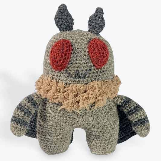 A crochet mothman is on a white background. Mothman has a grey head, body, and legs with black and grey striped arms, black antennae, black wings, a fuzzy brown collar, and big deep red eyes. Mothman's details include a sewn V pattern for texture on the body and legs. Mothman has a black stitched mouth that is a silly/creepy smile. 