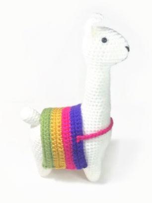 A white crocheted llama wearing a Sapphic Pride Flag striped blanket (purple, pink, yellow, green). The blanket is held on by a pink strap across the llama's chest. The llama has a black nose and black eyes. The llama's tail is white. The llama is standing on a white surface.